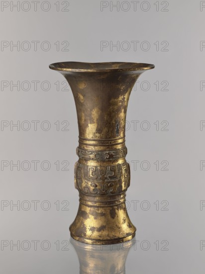 Zun vase, Chinese, ceremonial vessel vessel, taotie and beast motifs, meander and leiwen ornament, Qing dynasty 1644–1911, 19th century, China; height 31.7 cm, diameter 19.0 cm., A tall metal vessel with a flared rim and a narrower midsection sits against a plain light gray background, showing worn gold and dark brown patina across its surface; the midsection features a decorative band with carved, rounded motifs and raised linear rings above and below, and the base flares outward with similar worn gold and brown coloration, the vessel casting a faint reflection on a glossy surface below.