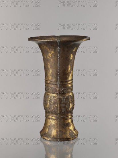 Zun vase; maker, Chinese; ceremonial vessel vessel, cup; taotie and fantastic beasts iconography; meander and leiwen ornament; Qing dynasty 1644–1911, 19th century ; China; height 31.7 cm, diameter 19.0 cm., A tall metallic vessel with a flared rim and a narrower waist, showing worn patches of gold and brown tones across its surface; a vertical seam runs down its center and there are decorative raised bands and a band of carved or embossed scroll-like patterns around the midsection; the metal shows areas of tarnish and patina in darker brown and greenish hues, and the piece sits on a reflective surface that mirrors its base; the visible colors include gold, brown, dark brown, greenish, and gray.
