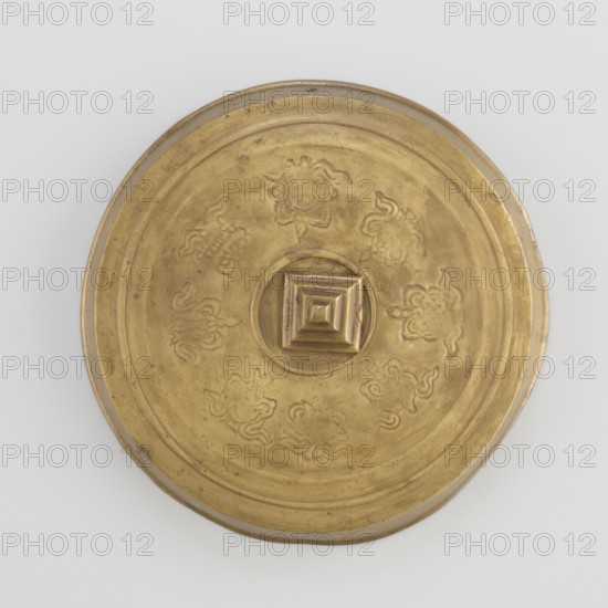 Cover; unknown creator role/craftsman; author; craft; Tibetan culture; unknown provenance; carved symbols of the Eight Preciousnesses ba bao; square shape with pyramidal boss; 19th century; Mongolia and Tibet place of origin; varia; diameter 12.7 cm, A round metallic object viewed from above with a raised central square knob and concentric ridges, showing embossed floral-like motifs arranged around the center; the surface appears worn and slightly uneven with some small scratches and patina. The visible colors are gold and muted brown on the metallic surface, with a light gray background.