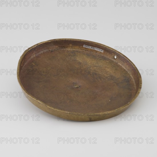 Cover; unknown creator/author/craftsman; Tibetan culture; unknown provenance; carved symbols of the Eight Preciousnesses Ba bao; square with pyramidal boss; 19th century; Mongolia and Tibet places of origin; varia; diameter 12.7 cm, A shallow round dish with a low rim, shown from an angled top view, made of metal with a worn, mottled brown and golden surface; the rim and outer edge show darker brown and greenish-golden tones, and there is a small irregular depression near the center and a tiny hole near the rim.