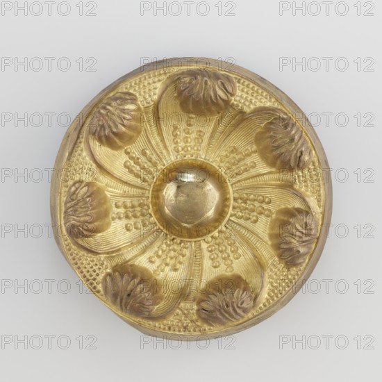 Seven-leaf rosette, craft, Tibetan culture, provenance unknown, rosettes, leaves ornament, decorative elements, 19th century, Tibet, Mongolia, varia; height 4.3 cm, diameter 12.4 cm, A circular gold-colored metal object with a raised central dome and a decorative floral-like pattern radiating outward, featuring a group of petal-shaped ridges and textured bead-like rows between them; the petals have darker brownish-gold shaded areas at their tips and along their edges, while the rest of the surface is bright yellow-gold with varying metallic highlights and subtle shadows against a pale neutral background.