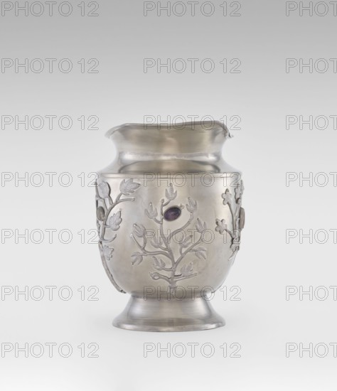 Jug, ornamented with plant motifs and fruit, featuring applied plastic decoration of flowering leafy bunches with oval set semi-precious stones; Chinese, Qing dynasty 1644–1911, 19th century; porcelain/ceramic vessel, height 20.6 cm, width 23.0 cm; subject includes plum trees; function: teapot/vessel., A metal vessel with a rounded body, a flared foot, and a wide, slightly undulating rim, shown against a light neutral background; the vessel has a smooth silvery surface with a subtle sheen and is decorated with raised botanical motifs of branches and leaves in the same metal, and a few oval dark stones set within the floral designs; overall colors visible include silver and a dark purplish tone in the inset stones, with soft gray gradients in the background.