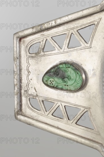 Platter, Chinese, openwork with bats and coins iconography, Qing dynasty 1644–1911, 19th century. Platter on a foot with two trapezoidal handles, vessel. Height 48.7 cm; width 24.2 cm., A close-up of a metallic object showing a section of its decorative surface in silver tones with areas of tarnish and darker discoloration; the design includes several triangular cutouts and scalloped engraved lines, with a green carved stone set into an oval bezel at the center that shows carved floral-like patterns and small perforations; the background is a smooth light gray.