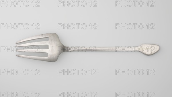 Serving fork; maker, Chinese; cutlery decorated with bats and coins; used in kitchens; Qing dynasty 1644–1911, 19th century; China; height 24.9 cm, width 4.9 cm., A single metal serving fork lies horizontally across a light gray background, showing a long thin handle that ends in a rounded, slightly teardrop-shaped grip and widens into a flat head with four broad, tapered tines separated by elongated slots; the metal surface appears worn with scuffs and scratches, and the visible colors are various shades of silver, gray, and off-white.