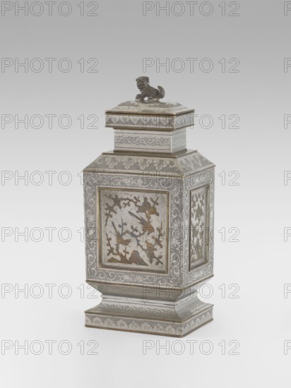 Teapot with a lid, Teapot with a profiled foot and neck, with a rectangular lid topped with a modeled lion figurine. Decorated with panels filled with a motif of a blossoming plum tree and birds, bordered with plant twigs panel motif engraved and set with metal. Foot, neck and lid decorated with plant ornament on a powdered background. Materials/style references: Chinese, huaniao style, Qing 1644–1911, 19th century. Iconography: lions, birds and flowers, fo, clouds, plant twig. Function: vessel teapot. Dimensions: height 27.3 cm, width 11.5 cm, depth 8.3 cm., A decorative metal container with a square, tapered body and a matching lid topped by a small sculpted animal figure, shown against a light gray background. The container has ornate engraved and inlaid panels on each side featuring floral and bird motifs, with borders of scrolling foliage and patterned bands. The lid and stepped base carry similar engraved patterns and flourishes. Visible colors include silver, gray, and accents o...
