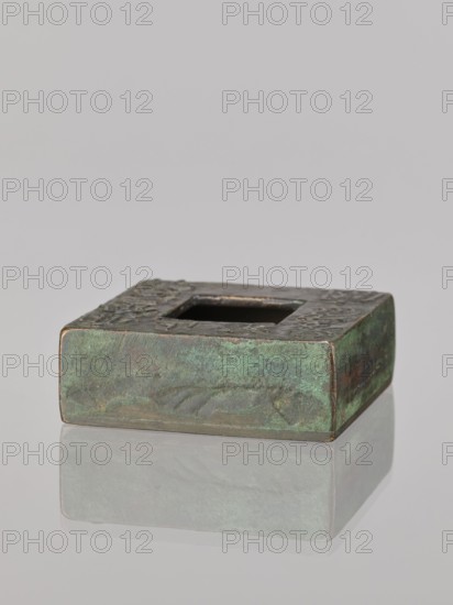 Water container, Narozno Eckersdorf; Chinese art, Chinese culture; inscriptions Chinese; Qing 1644–1911, 19th/20th century, China; vessel; height 2.6 cm, width 7.5 cm, depth 7.4 cm., A low square metal object with a square central hole sits on a reflective surface, showing a worn patina of green and brown with areas of darker bronze, and the top edge has textured raised decoration; the reflection beneath repeats the green and brown tones against a neutral light gray background.