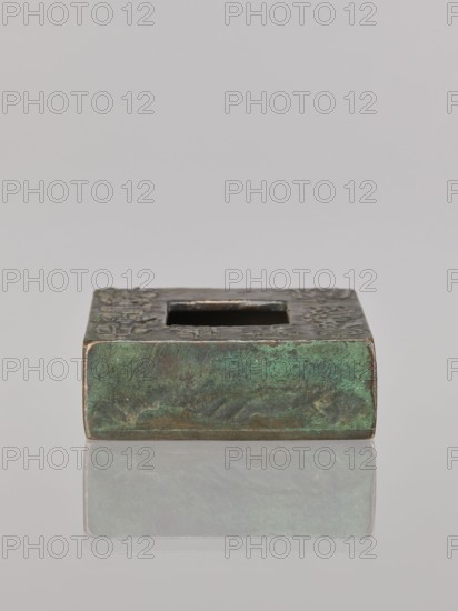 Water container, Narozno Eckersdorf, Chinese art, Chinese culture, inscriptions Chinese, restitution provenance, Qing 1644–1911, 19th/20th century, China, vessel, height 2.6 cm, width 7.5 cm, depth 7.4 cm, A small rectangular metallic object with a square hole in its top, resting on a reflective surface; the metal shows a mottled green patina with areas of darker and lighter green, brownish and brassy tones, and some worn edges where a warmer metallic color is visible, and the background is a pale gray.