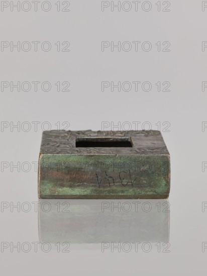 Water container, Narozno Eckersdorf, Chinese art, Chinese culture, Chinese inscriptions, restitution provenance, Qing 1644–1911, 19th/20th century, China, vessel, height 2.6 cm, width 7.5 cm, depth 7.4 cm, A rectangular metal block with a square opening on its top surface, showing a patina of green and brown tones with areas of darker gray and hints of reddish-brown, resting on a reflective light gray surface that produces a soft mirrored reflection beneath it.