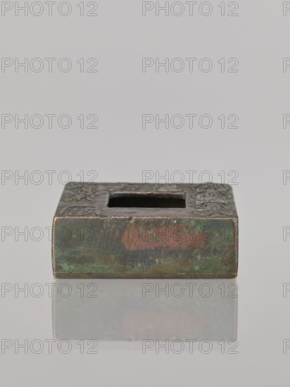Water container, Narozno Eckersdorf — Chinese art; Chinese culture; Chinese inscriptions; provenance noted; Qing dynasty 1644–1911, 19th–20th century; China place of origin; vessel; height 2.6 cm, width 7.5 cm, depth 7.4 cm, A small square metal object with a central square opening sits on a reflective surface, showing a faint mirror image below. The object has an aged patina with visible green and brown tones, areas of reddish discoloration, and darker grayish sections. The top surface appears textured with raised decorative elements and worn edges, and the background is a uniform light gray.