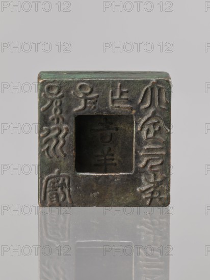 Water container, Narozno Eckersdorf — Chinese art, Chinese culture, inscriptions Chinese; Qing 1644–1911, 19th/20th century, China; vessel; height 2.6 cm, width 7.5 cm, depth 7.4 cm, A square, hollow metal block with a central square opening sits upright on a reflective surface, showing a weathered, uneven surface with raised decorative patterns and incised shapes around the opening; the metal displays greenish patina, dark brown, and bronze tones, with areas of lighter worn highlights and subtle variations in those colors, and the reflection beneath mirrors the block’s shape and textures.