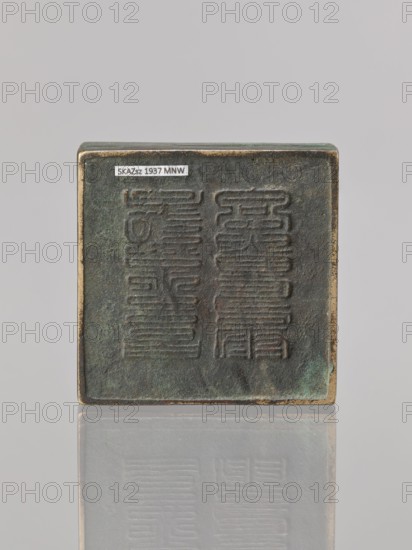Water container, creator unknown, Narozno Eckersdorf; Chinese art; Chinese culture; Chinese inscriptions; Qing dynasty 1644–1911; 19th–20th century; China; vessel; height 2.6 cm, width 7.5 cm, depth 7.4 cm., A square, flat object with a worn, textured surface in dark green and brown tones sits upright against a light gray background, showing raised, rectangular patterns across its face; the object's edges reveal a metallic, slightly tarnished rim in brass-like color, and a faint reflection of the object appears below on a glossy surface.