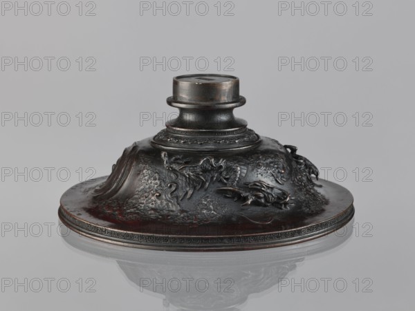 Cover, Chinese art, Chinese culture, Qing 1644–1911, meander ornament, dragons iconography, 19th century, China, varia, height 10.2 cm, diameter 23.8 cm, A low, rounded metallic object with a circular base and a short cylindrical neck sits on a reflective surface; the object has sculpted, raised decorative reliefs resembling flowing shapes and curled forms around its sides, with textured, irregular surface details. The finish shows dark brown and black tones with areas of reddish-brown highlights and subtle metallic sheen, and the reflective surface beneath shows a muted mirror image of the object. The overall background is a uniform light gray.
