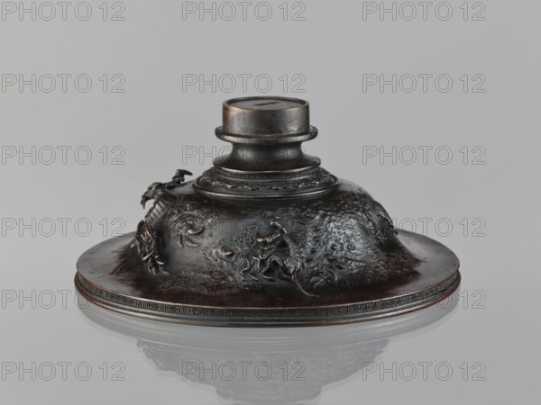 Cover, repossession Salzburg, Chinese art, Chinese culture, Qing 1644–1911, meander ornament, dragons iconography, 19th century, China, varia, height 10.2 cm, diameter 23.8 cm, A dark metallic rounded object with a raised central cylindrical neck sits on a shallow circular base, showing a textured surface with sculpted relief forms resembling curved organic shapes and small protrusions; the metal appears dark brown to black with subtle reddish-brown highlights and areas of lighter metallic sheen, and the base edge has a patterned band in similar dark tones, all set against a light gray background with a soft reflection beneath the object.