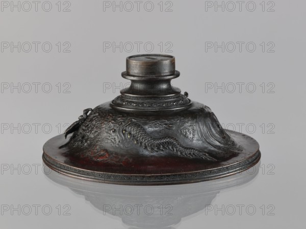 Cover, repossession Salzburg, Chinese art, Chinese culture, Qing 1644–1911, meander ornament, dragons iconography, 19th century, China, varia, height 10.2 cm, diameter 23.8 cm, A low, rounded metal object with a central cylindrical neck and a wide base sits on a reflective surface, showing a mirrored underside; the piece is dark brown to almost black with areas of reddish-brown patina and faint gold highlights; the base rim has a decorative band and the sloped body features raised, textured relief elements including a scaly, sinuous creature and flowing wave-like forms; the cylindrical neck has layered rings and a slightly lighter, worn top; the background is a smooth, pale gray.