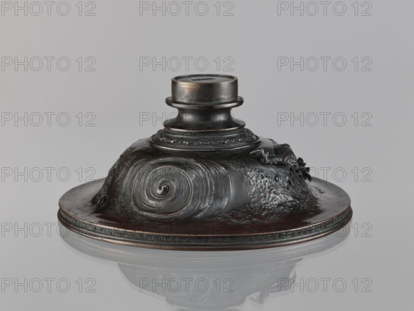 Cover, role: craftsman, personal entries, repossession Salzburg, Chinese art, Chinese culture, Qing 1644–1911, meander ornament, dragons iconography, 19th century, China, varia, height 10.2 cm, diameter 23.8 cm, A dark metallic object with a low, wide base and a raised central neck sits on a reflective surface, showing colors of dark brown, black, and subtle bronze highlights, with a smooth spiral motif on one side and rough textured areas on another, and a small sculpted element attached near the top rim; the base edge features a repeated decorative band and the object casts a soft reflection on a pale gray background.
