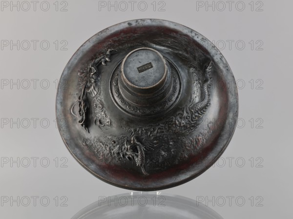 Cover, role/classification of the creator/craftsman unspecified, personal entries, Chinese art; China, Qing dynasty 1644–1911, 19th century; meander ornament, dragons iconography; varia; height 10.2 cm, diameter 23.8 cm., A round metal object viewed from above with a raised central cylindrical section and a flared rim, featuring sculpted relief decorations including dragon-like creatures and textured wave-like patterns; the surface shows a patina with dark gray, black, and reddish-brown tones, and areas of lighter gray highlights, sitting on a reflective neutral background that appears gray.