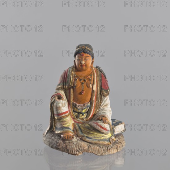 Figurine of a seated man, Chinese, ceramics, male clothing, seated figure, male figurine, 19th century, China, sculpture, figurine, height 15 cm, A painted figurine of a seated person wearing layered robes in colors including red, white, yellow, green, blue, and brown, with a dark bluish hair bun and a reddish dot on the forehead, a beaded necklace with small hanging ornaments, one hand resting on a knee and the other draped over a knee, bare toes visible, seated on a textured brown base with a rectangular object beside them, all shown against a plain light gray background.