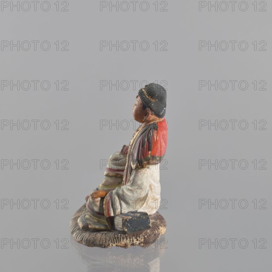 Figurine of a seated man, Chinese, Chinese art, ceramics, male clothing, seated figure, male figurine, 19th century, China, sculpture, figurine, height 15 cm, A small painted figurine of a seated person shown in profile on a circular textured base, featuring colors including gray, red, black, white, brown, beige, yellow, and a hint of green; the figure wears a black head covering with gold accents, a red garment over the shoulders, and a mostly white robe with colored trim, with visible worn and chipped paint and a few areas of darker weathering on the base and clothing.