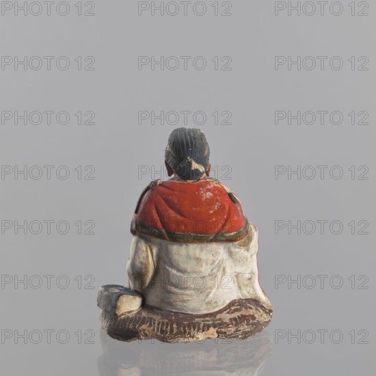 Figurine of a seated man, Chinese, ceramics, male clothing, seated figure, male figurine, 19th century, China, sculpture, figurine, height 15 cm, A small painted figurine shown from behind sitting on a textured base, with dark hair tied back, a red upper garment with worn areas, a light-colored lower garment with visible creases and weathering, and brown and beige tones on the base and edges; the background is a uniform pale gray.