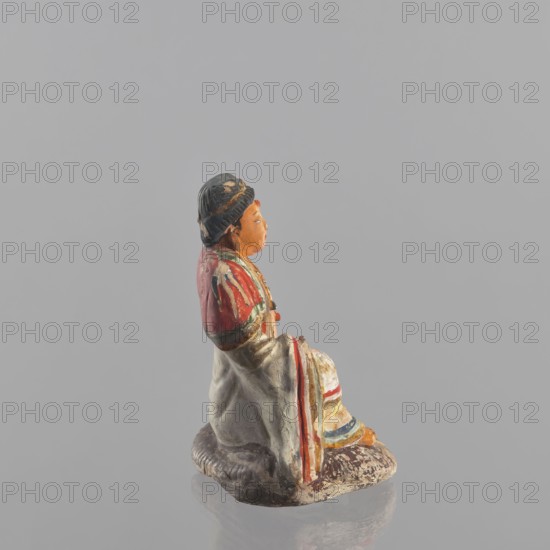 Figurine of a seated man; creator, Chinese; Chinese art; ceramics; male clothing; seated figure; male figurine; provenance, 19th century : China; sculpture; figurine; height 15 cm, A small painted figurine shown in profile, featuring a seated figure with a dark blue hat and orange-toned face, wearing a red upper garment with green and white trim and a long off-white robe draped over the legs; the robe has visible stripes of red, blue, and yellow near the hem and sits on a textured brown base, all set against a smooth light gray background.