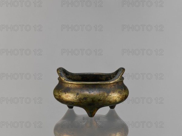 Li-type incense burner, Chinese, ceremonial Li-type ritual vessel, Ming style, Qing 1644–1911, backdated signatures, 19th century, China, vessel; height 6.8 cm, diameter 12.9 cm, A small metal vessel with a rounded body and a short flared rim sits centered on a reflective surface, showing a partial reflection beneath it; the vessel has a patinated surface with mixed tones of gold, dark brown, and greenish tarnish, and it rests on three short pointed feet with a slightly worn and mottled texture, while the background is a smooth, even gray.