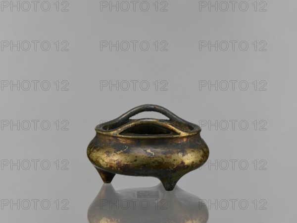 Li-type incense burner, Chinese, ceremonial Li-type ritual vessel, Ming style, Qing 1644–1911, backdated signatures, 19th century, China; vessel; height 6.8 cm, diameter 12.9 cm, A small metal vessel with a rounded body and a pair of curved handles across its open top sits on a reflective surface, showing a mirrored underside; the vessel has an aged, mottled finish with areas of gold, dark brown, and greenish patina, and it stands on three short tapered legs against a plain gray background.