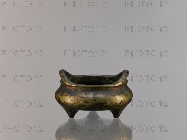 Li-type incense burner, Chinese, ceremonial ritual vessel in Li-type form, Ming style, Qing 1644–1911, possibly 19th century, backdated signatures, China; vessel; height 6.8 cm, diameter 12.9 cm, A small, low metal vessel with a rounded body and a flared rim supported on three short legs sits centered against a plain, light gray background, the metal surface showing a mottled patina of gold, green, brown, and dark gray with areas of darker discoloration and subtle reflective highlights, and the object’s reflection visible on the smooth surface beneath it.