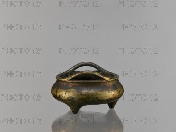 Li-type incense burner, Chinese, ceremonial/ritual vessel in Li-type form, Ming style, Qing 1644–1911, possibly 19th century, backdated signatures, China; vessel, height 6.8 cm, diameter 12.9 cm., A small metallic vessel with a rounded, bulbous body and three short tapered legs sits centered against a smooth light gray background, its surface showing a mottled patina of brassy gold, dark brown, and greenish tones; the vessel has a thick rim and an arched double handle spanning the opening, with darker edges and worn areas that reflect subtle highlights, and a faint reflection of the object appears on the glossy surface beneath it.