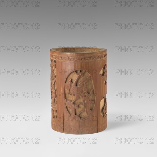 Brush vessel, Chinese, Qing dynasty 1644–1911, early 20th century. Porcelain container with bas-relief decoration of figural scenes in a landscape and images of birds; reserves with bas-relief decoration of various shapes. Height 16.4 cm; diameter 11.9 cm., A cylindrical wooden container with a warm brown patina stands upright on a pale surface against a neutral gray background, its exterior carved with an oval relief on the front depicting a stylized bird with outstretched wings and flowing tail, and flanked by carved openwork panels showing organic and geometric motifs; the rim shows a carved band with repeating leaf-like shapes, and the wood grain and some darker and lighter brown variations are visible across the piece.