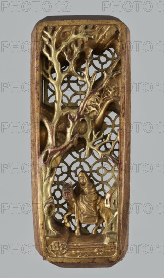 Monk on Horseback Under a Tree — decorative panel;, Chinese; horses and monks iconography; geometric motifs; Qing dynasty 1644–1911; 19th century; carved sculpture in the shape of an elongated rectangle with openwork geometric background; dimensions: height 4.1 cm, width 27.3 cm, depth 10.7 cm; provenance unknown., A vertical carved panel with a rectangular frame showing a detailed scene in gold and brown tones, featuring a group of leafless branching forms and textured surfaces, a lattice-like patterned background visible through openings, and a small hooded figure seated on a four-legged animal near the lower center; the carving shows traces of red accents and the frame edges and background have wood-colored brown areas against a neutral gray backdrop.