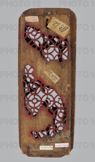 Monk on Horseback Under a Tree — decorative panel;, Chinese; horses and monks iconography; geometric motifs; Qing dynasty 1644–1911, 19th century; carved sculpture in the shape of an elongated rectangle with openwork background of geometric ornament; materials not specified; dimensions: height 4.1 cm, width 27.3 cm, depth 10.7 cm; provenance unknown., A vertical wooden panel with a rectangular shape and rounded top and bottom edges showing a carved lattice of interlocking circular floral patterns revealing a background through irregular openings, the carved areas edged with worn red paint and the wood surface various shades of brown with darker worn spots and scratches, a few small paper labels affixed to the surface but not described in their markings, and a pale gray background behind and visible through the carved openings.