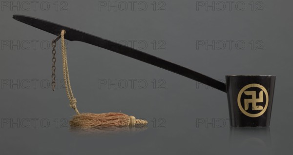 Ritual ladle, Chinese, Qing 1644–1911, Chinese Buddhism ritual vessel with swastikas painted gold. A chain and silk cord with a tassel hang from the handle. Late 19th century, China. Length 57.5 cm., A long black ladle-like object with a glossy finish extends horizontally across the image, attached at the right end to a short black cup also with a glossy surface; a circular decorative motif appears on the cup in a contrasting gold color. From near the left end of the handle hangs a short chain and a braided beige cord that ends in a tassel made of coarse brown and tan fibers; the tassel rests on a reflective grey surface that mirrors the objects. The background is a smooth, uniform grey.
