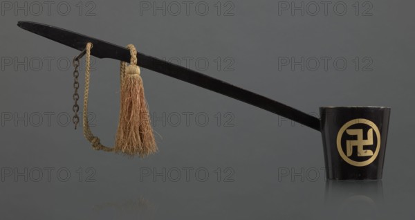 Ritual ladle, Chinese, Qing dynasty 1644–1911, late 19th century. Buddhism ritual vessel with swastikas painted gold. A chain and silk cord with a tassel hang from the handle. Length 57.5 cm., A long black ladle with a tapered handle extends across the image, ending in a cup-shaped bowl that is black with a circular lighter-colored emblem on its side; a braided tan cord and a frayed tan tassel hang from the handle along with a short dark metal chain, and the background is a smooth gray.