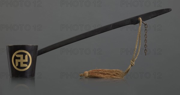 Ritual ladle, Chinese, Qing dynasty 1644–1911, late 19th century. Material/subject: ritual vessel associated with Chinese Buddhism; swastikas painted gold. A chain and silk cord with a tassel hang from the handle. Length: 57.5 cm., A long black ladle-like object with a straight handle stretches across a gray background, attached at one end to a small black cup; the cup has a circular pale yellow design on its side. From the handle near the far end hangs a braided beige cord and a short rusty metal chain, the cord tied and ending in a frayed tan-brown tassel that lies on the reflective surface. The image shows colors including black, gray, beige, tan-brown, rusty brown, and pale yellow.