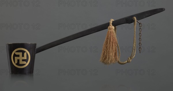 Ritual ladle, Chinese, Qing 1644–1911, Chinese Buddhism, ritual vessel with swastikas painted gold. A chain and silk cord with a tassel hang from the handle. Late 19th century, China. Length 57.5 cm., A long black ladle-like object extends across the image, attached at one end to a short black cup with a circular pale yellow decorative motif on its side, and at the other end to a braided tan cord that holds a tassel of coarse light brown fibers and a short rusty metal chain; the background is a uniform dark gray and the surface beneath the objects reflects them faintly.