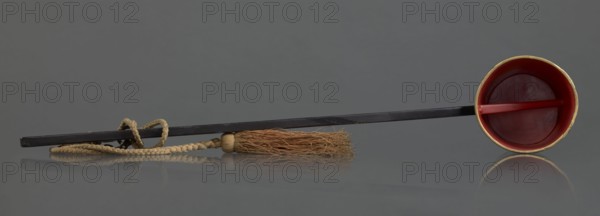 Ritual ladle, Chinese, Qing dynasty 1644–1911, late 19th century. Buddhism ritual vessel, painted gold swastikas; a chain and silk cord with a tassel hang from the handle. Length 57.5 cm., A long dark rod lies horizontally across a smooth gray surface with its reflection visible, attached near one end to a braided beige cord that forms a loop and leads to a tassel of coarse light brown fibers bound by a small beige cap; at the opposite end the rod is fitted into a shallow round cup that is red inside with a thin golden rim and a short red crossbar across its interior.
