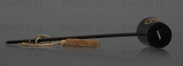 Ritual ladle, Chinese, Qing dynasty 1644–1911, Chinese Buddhism ritual vessel, late 19th century, China; materials: chain and silk cord with a tassel hang from the handle; swastikas painted gold; length 57.5 cm., A long dark rod with a cylindrical dark head lies horizontally on a reflective gray surface against a gray background, a twisted tan rope looped around the rod and attached to a beige tassel with frayed tan and light brown fibers, and the cylindrical head shows a curved gold pattern on its side; visible colors include dark brown or black, gray, tan, beige, light brown, gold, and a small area of white.