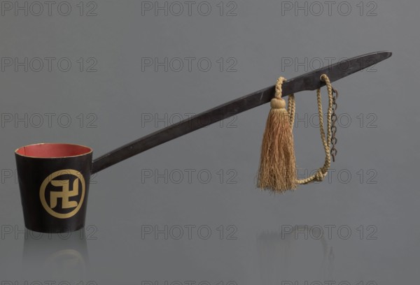 Ritual ladle; maker, Chinese; Qing dynasty 1644–1911; Chinese Buddhism ritual vessel; late 19th century, China; materials: chain and silk cord with a tassel hang from the handle; swastikas painted gold; length 57.5 cm., A long dark handle attached to a dark cup with a red interior rests against a gray background, with a gold-colored circular emblem on the cup's exterior partially visible; a braided tan cord with a tassel of natural brown fibers hangs near the handle's end alongside a short dark metal chain, and the scene reflects faintly on the surface below.