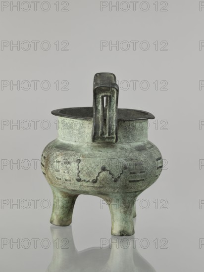 Ding-type incense burner, maker, purchase provenance. Chinese; ceremonial vessel with geometric ornament; associated with Chinese philosophy and fortune telling. Qing dynasty 1644–1911, 17th–18th century . China. Vessel; height 23 cm, width 19.4 cm, diameter 18.7 cm., A greenish bronze tripod vessel with a rounded body and a flared rim standing on three stout legs, showing a mottled patina of green and darker brown; the body is decorated with incised geometric patterns and raised dot-and-line motifs, and a rectangular loop handle rises from the rim with a darker, more worn surface; the vessel casts a soft reflection on a pale gray surface beneath it against a neutral light gray background.