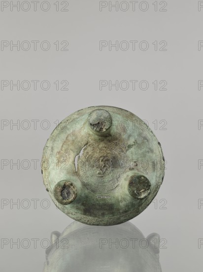 Ding-type incense burner, maker, Chinese; ceremonial vessel; geometric ornament; associated with Chinese philosophy and fortune telling; Qing dynasty 1644–1911, 17th–18th century, China; height 23 cm, width 19.4 cm, diameter 18.7 cm, A round metal object with a weathered, patinated surface in shades of green and brown sits centered against a neutral gray background, showing a circular top with three raised circular bosses and areas of corrosion and rough texture; the metal displays mottled green, bluish-green, brown, and darker gray-green tones with some lighter green highlights, and the object casts a soft reflection below on a glossy surface.