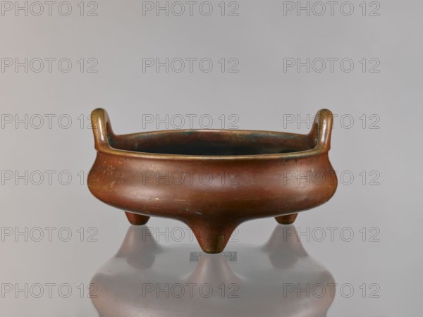 Li-type incense burner, Chinese, ceremonial/ritual vessel. Ming style Ming 1368–1644; Qing 1644–1911; Xuande 1426–1435. Antidated signatures. Cast in golden-red bronze with a light reddish-brown patina. Flattened low body, twig-shaped handles, low legs. Signed. 18th century ; China. Vessel censer. Height 13 cm; diameter 26 cm; depth 6.5 cm., A low, round three-legged vessel with two short upward-curving handles, showing a smooth patinated surface in brown and bronze tones with areas of greenish verdigris, a darker interior, and a soft reflection of the object on a pale gray surface against a light gray background.