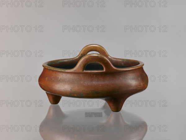 Li-type incense burner, Chinese, Ming dynasty style Ming 1368–1644; associated with Qing 1644–1911 and Xuande period 1426–1435. Cast in golden-red bronze with a light reddish-brown patina; flattened low body, twig-shaped handles, low legs. Antidated signatures; signed. Ceremonial ritual vessel censer, 18th century, height 13 cm, diameter 26 cm, depth 6.5 cm., An antique three-legged bronze vessel with a rounded body and a smooth rim sits on a reflective surface, showing a subtle mirrored reflection beneath it; the vessel has a pair of arching handles rising from opposite sides and an interior that appears darker than the exterior; the surface shows mottled patina with visible spots of green and darker brown on a dominant reddish-brown bronze color, and the background is a soft, neutral gray.