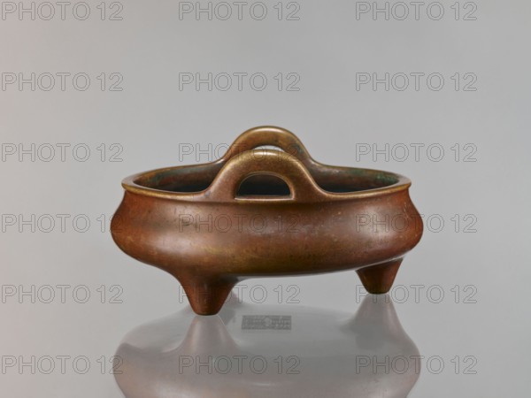 Li-type incense burner, Chinese, cast in golden-red bronze with light reddish-brown patina. Flattened low body with twig-shaped handles and low legs. Inscribed/antidated signatures; style associated with Ming 1368–1644, Xuande 1426–1435 and Qing 1644–1911. Function: ritual/ceremonial censer. 18th century, China. Height 13 cm; diameter 26 cm; depth 6.5 cm., A low, rounded bronze vessel with a warm reddish-brown patina sits on a reflective surface; it has a wide rim, a pair of rounded arching handles with cut-out centers, and three short conical legs. The vessel shows areas of darker brown and subtle greenish corrosion inside the rim and along the interior, with small variations in tone and mottling across the exterior. The background is a smooth light gray, and the reflection of the vessel and its legs appears muted on the glossy surface beneath.