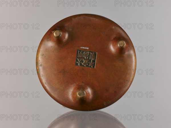 Li-type incense burner, Chinese, ceremony vessel in Ming/Qing styles Ming 1368–1644; Qing 1644–1911; Xuande period 1426–1435 antidated signature. Cast in golden-red bronze with light reddish-brown patina. Flattened body with twig-shaped handles and low legs. Signed. 18th century, China. Vessel censer. Height 13 cm; diameter 26 cm; depth 6.5 cm., A round, flat object with a mostly reddish-brown metal surface and a patina showing areas of darker brown and small greenish spots; the object has three rounded protrusions each topped with a brighter gold-toned circular cap and a recessed square panel in the center with carved linear patterns; the item rests on a reflective gray surface that creates a faint mirrored semicircle beneath it, and the background is a smooth light gray.