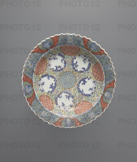 Plate, Colored porcelain, decorative vessel with serrated edge. Decoration: in the mirror, round reserves filled with plant and geometric ornament; background of small, densely covering plant ornament; on the reverse, on the rim, a motif of seedlings chidori flying over sea waves. Japanese, Arita, c. 1800–1860. Vessel height 5.5 cm, width 27.6 cm., A decorative round ceramic plate with a scalloped edge resting against a solid gray background, featuring an intricate multicolored pattern; the plate shows a central circular medallion with a repeating geometric motif surrounded by a ring of floral and scrollwork designs in blue, green, gold, and pale yellow, interspersed with several white cartouche-like areas containing blue floral silhouettes, and framed by alternating red, orange, and blue patterned panels around the rim, with accents of light turquoise, beige, and touches of brown.