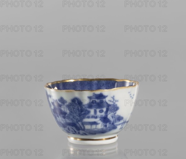 Bowl, ceramics, blue-and-white porcelain, landscape motifs, Qing style, 19th century Qing dynasty, 1644–1912, China; part of a 22-piece tea and coffee set teapot, 6 cups, 6 saucers, a bowl, a cover, and a plate; vessel; height 5 cm, diameter 8 cm., A small ceramic cup with a scalloped rim and a gold trim sits on a reflective surface against a neutral gray background. The exterior of the cup is white with a blue painted landscape scene showing stylized trees and architectural shapes, and the interior appears blue with a subtle patterned texture. The rim and the base have gold accents, and the overall colors visible are white, blue, gold, and gray.
