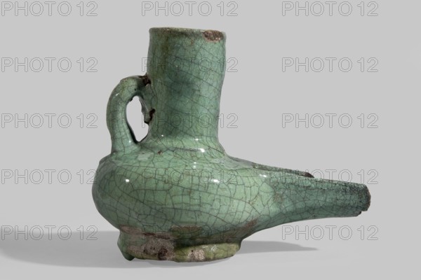 Jug-shaped oil lamp with a handle at the neck and a long burner, covered with a turquoise glaze on brown clay; ring foot. Glazed utility ceramic, Egyptian, Mamluk Sultanate 13th–14th century. Arab/Mamluk style. Vessel; weight 0.38 kg; height 11.0 cm; width 13.4 cm., A ceramic vessel with a rounded body, a tall cylindrical neck, a short handle connecting neck and shoulder, and an elongated spout extending horizontally; the surface shows a crackled glaze with a green tone and darker crack lines, with areas of darker brownish discoloration and small patches of unglazed rough material, all set against a neutral gray background with a soft shadow beneath.