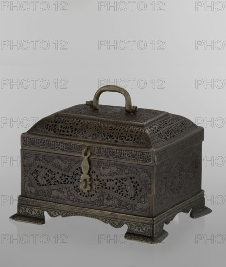 Box; rectangular on four feet with a lid and a handle on top. Persian Iran, Qajar dynasty, 19th century. Materials and exact technique not specified. Decorated with animals, hares, fantasy figures, hunting scenes, archers, handles, figurative motifs, and inscriptions. Height 19.9 cm; width 15.6 cm; depth 13.8 cm., An ornately carved metal box with a domed lid and a rounded handle on top sits on a reflective surface; the box is dark bronze with areas of lighter brass tones, showing intricate openwork panels and decorative relief patterns on all visible faces, a hinged clasp at the front, raised feet with engraved borders, and a series of repeating geometric and floral motifs across the lid and body.