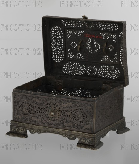 Box; Persian Qajar dynasty, 19th century; Iran. Rectangular box on four feet with lid and top handle. Decorated with figurative motifs including hares, fantasy figures, hunting scenes, archers, animals, inscriptions and handles. Materials and exact, Dimensions: height 19.9 cm, width 15.6 cm, depth 13.8 cm., A decorative metal box with an open lid showing intricate perforated patterns; the box has a dark metallic surface with areas of lighter metallic highlights and a few greenish patina spots. The lid features a repeating pierced design and two small metal hooks, and the interior shows similar pierced patterns in dark metal. The exterior sides display detailed engraved and relief ornamentation with flowing vegetal and geometric motifs in dark metal tones with subtle lighter edges. The box rests on a raised ornate base with carved feet and patterned borders in matching dark metallic colors against a pale gray background.