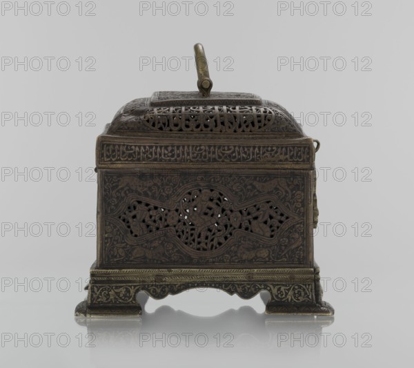 Box;, gift; Islamic art, Persian Qajar dynasty, 1785–1925; 19th century, Iran. Rectangular box on four feet with a lid and a handle on top. Decorated with animals, hares, fantasy figures, hunters/archers, handles, figurative motifs; inscriptions. Height 19.9 cm, width 15.6 cm, depth 13.8 cm., A small ornate metal box with a hinged domed lid and a carry handle, shown from the front on a reflective surface. The box has intricate pierced and engraved patterns across its surfaces, including decorative openwork panels on the front and top. The metal appears in shades of dark brown and aged brass, with darker recessed areas and lighter brass highlights along edges and raised details. The base rests on short ornate feet with scrolling and floral motifs, and the lid has a rectangular raised section with more pierced decoration. The background is a smooth light gray.
